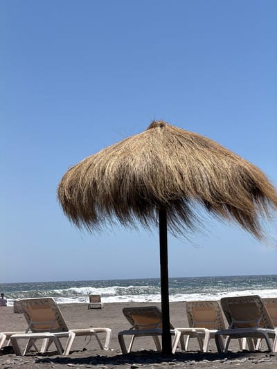 A beach umbrella with a straw-like covering stands beside lounge chairs on a beach with dark sand. The ocean waves are visible alongside the chairs. The sky above is a clear blue. The umbrella and chairs are arranged in a line next to the water. The image is oriented vertically, emphasizing the height of the umbrella.