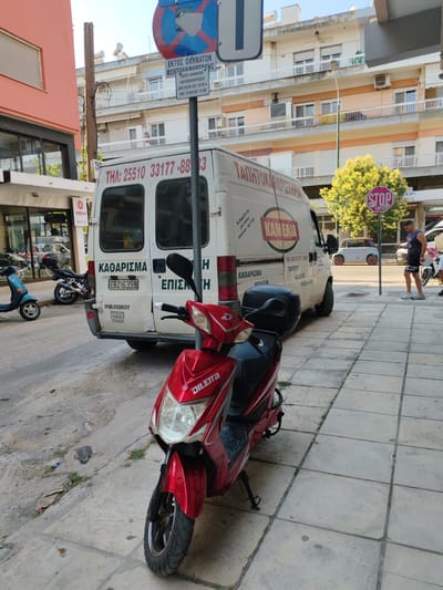 A street scene in Alexandroupolis, Greece, features a red Diletto scooter parked on a sidewalk next to a white Peugeot van. The van has the text "ΚΑΘΑΡΙΣΜΑ" and a phone number "ΤΗΛ: 25510 33177-88303" on its side, indicating a cleaning service. There are other motorcycles and a blue scooter parked nearby. A man in athletic wear is visible in the background. Traffic signs, including a "STOP" sign and a no-parking sign, are present. Buildings with balconies line the street. The environment appears to be a commercial or residential area with trees and overhead wires. The ground is paved with concrete and tiles.