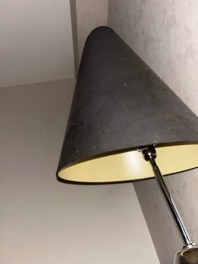The image shows a close-up of a dark lampshade with a yellow interior, partially obscuring a textured wall. The lampshade has a conical shape and appears to be made of fabric. Small specks, possibly dust or debris, are visible on the exterior of the lampshade. A chrome lamp stem is visible extending from beneath the shade. The wall behind the lamp has a light, textured finish, with some areas appearing darker than others, suggesting shadows or variations in the material. The ceiling area is visible in the upper left corner, appearing to be a plain, light color, meeting the wall at an angle. No people or specific geographical markers for Chantepie, France are discernible in this image.