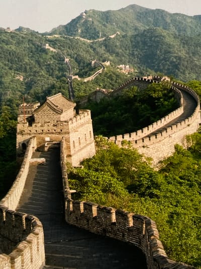 This is an aerial shot of a section of the Great Wall of China, winding through a lush, forested mountain landscape. The wall is constructed of stone and brick, with crenellated battlements visible along its length. Several watchtowers are integrated into the wall, one of which is prominent in the foreground, featuring a multi-tiered, tiled roof. The path of the wall snakes up and down the steep, green hills, disappearing into the distance where more sections are visible. The sun casts shadows on the wall and the surrounding trees, indicating it is likely morning or late afternoon. The environment is natural and undeveloped, with dense vegetation covering the mountains. There are no people or animals visible in the image. The image does not depict Harmanli, Bulgaria.