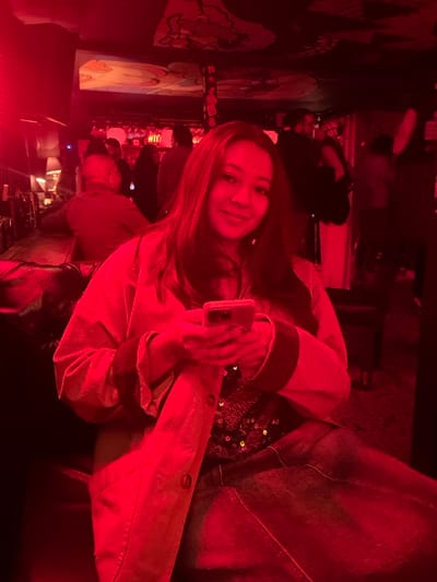 The image portrays an indoor scene, consistent with a bar or lounge environment, illuminated predominantly by an intense red ambient light source. A female individual is seated in the foreground, positioned centrally, facing directly towards the viewer with a faint smile. She has long, dark hair and is dressed in a light-colored, long-sleeved outerwear, potentially a jacket or coat, over a dark, sequined top, and dark trousers. She holds a smartphone with both hands, appearing to interact with it; the device exhibits multiple rear camera lenses. Her fingernails are painted a dark color. To the left of the primary subject, a dark bar counter is visible, along with a small, shaded table lamp and two male patrons seated with their backs partially to the camera. The background features several additional blurred figures, some standing, indicative of a crowded social gathering. The ceiling above displays abstract or decorative artistic elements. The scene is located within the city of Bogota, Capital District, Colombia; however, the specific venue or neighborhood cannot be determined from the visual information provided.