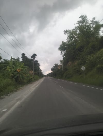 The image depicts a cloudy, overcast day along a paved road in Seuneubok, Indonesia. The perspective is from inside a vehicle, looking forward. The road is flanked by lush green vegetation and trees on both sides. On the left side of the road, there is a yellow diamond-shaped warning sign depicting a steep incline or decline. Utility poles and power lines run alongside the road. In the distance, several vehicles are visible on the road. The overall scene suggests a rural or semi-rural setting.