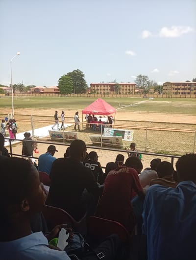 The image depicts a scene at what appears to be a sports stadium in Isasi, Nigeria. The foreground shows a section of seated spectators, most of whom are facing away from the camera. The middle ground features a running track and a grass field. A red tent is set up on the track, with people standing around it. A white banner or platform is placed in front of the tent, with logos and lettering visible. A wire fence separates the spectators from the track area. In the background, there are several multi-story buildings and trees. The sky is light blue with a few scattered clouds. A streetlight is visible on the left side of the field. People are walking on the track near the tent.