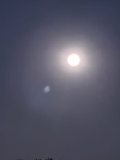 The image depicts a nighttime sky over Passignano sul Trasimeno, Italy. A bright, circular celestial body, likely the moon, is prominently positioned in the upper right quadrant of the image. Surrounding the moon is a halo of light, suggesting atmospheric conditions or lens flare. To the upper right of the moon, a faint, small, white speck is visible, possibly a star or distant light source. A hazy, oval-shaped blur of light is located in the middle left of the image. The sky is a gradient of dark blue and grey, with a darker, silhouetted treeline visible at the very bottom of the frame. The overall scene is dark and atmospheric.