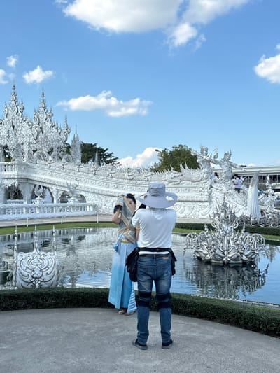 The image depicts an outdoor scene at Wat Rong Khun, commonly known as the White Temple, in Ban Rong Khun, Chiang Rai, Thailand. A bright blue sky with scattered white cumulus clouds dominates the upper half of the frame. The central and background elements consist of the highly ornate, pristine white architectural structures of the temple, featuring intricate carvings, multiple spires, and mythological figures, partially reflected in a calm body of water in the middle ground. Green trees and foliage are visible behind the white structures.
In the foreground, two individuals are present on a concrete path adjacent to the water. A person positioned with their back to the viewer, wearing a light grey broad-brimmed hat, a white short-sleeved shirt, blue jeans with black knee patches, and dark athletic shoes, holds a camera or smartphone to their eye, appearing to photograph the other person. The second individual stands to the left, facing partially away from the camera, dressed in traditional Thai attire consisting of a light blue gradient sarong-style lower garment and a light brown or gold upper garment or shawl, adorned with gold accessories. They are in a posed stance, suggesting they are being photographed. Additional unidentifiable individuals are visible in the background on the temple structures.