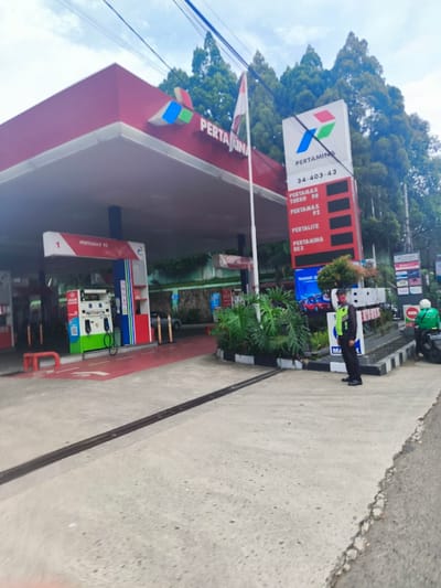 The image shows a gas station in Lembang, Indonesia, located at coordinates -6.832 latitude and 107.606 longitude. The gas station is a red and white structure with a canopy extending over the pumps. There is a sign on the side of the structure with the words "Pertamina" and a smaller sign with the words "Pertamax Turbo," "Pertamax," "Pertamax 92," "Pertalite," and "Pertamina Dex." A man in a black uniform is standing to the right of the sign, facing the camera. There is a single gas pump in the frame, and the ground is a red-colored concrete. The gas station is situated in a parking lot and bordered by greenery. A black motorcycle is parked next to a man in a green shirt. The sky is overcast.