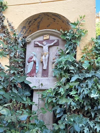 The image shows a religious scene depicted in stone relief within a niche on a yellow exterior wall. The relief features a depiction of Jesus Christ on the cross, with another figure, possibly Mary, depicted below. The scene is partially obscured by climbing ivy. The location is Bernkastel-Kues, Germany.