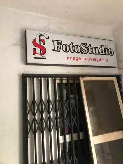 A vertical sign reads "FotoStudio...image is everything" with a stylized red "S" beneath. Adjacent to it is a window with a white frame, partially obscured by a black, expandable metal security grille. The wall appears aged and textured.