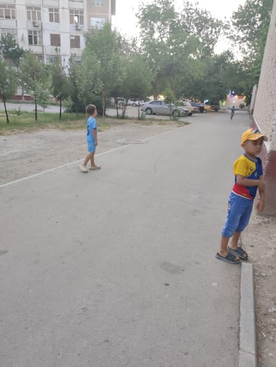 The media file shows two young boys standing on a paved walkway next to a building. The boy on the left wears a light blue t-shirt and shorts with white sandals. The boy on the right wears a yellow, blue, and red t-shirt with blue shorts and dark grey slides. He also wears a yellow baseball cap. They appear to be in an urban setting, with a building visible in the background. There are trees and parked cars in the distance. The light suggests it is either early morning or late afternoon, likely dusk. The scene is calm, with no particular activity taking place. The building appears to be an apartment complex with multiple windows. The paved walkway runs along the side of a sandy area. The overall impression is a quiet, everyday scene in a residential area.