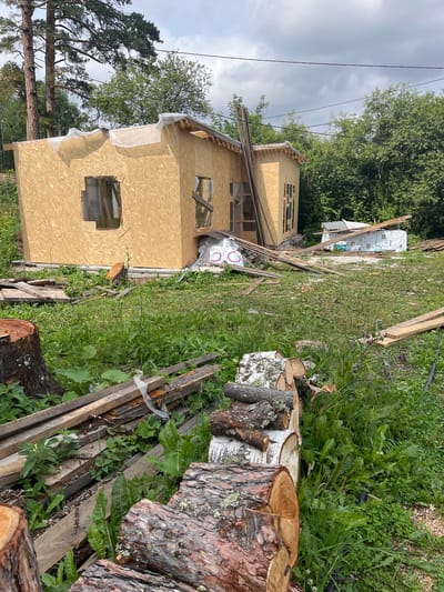 The image depicts a partially constructed wooden structure in what appears to be a rural or semi-rural setting. The building is framed with oriented strand board (OSB) and has openings for windows, some of which are covered with plastic sheeting. Construction materials, including lumber and bundles of what look like insulation or roofing material, are scattered around the site. In the foreground, several tree stumps and logs are visible, suggesting recent logging activity or preparation for firewood. The ground is covered with green grass and other vegetation. The sky is overcast with a mix of clouds. The presence of tall trees in the background indicates a wooded environment. The scene suggests an ongoing construction project in an outdoor, natural setting. The location is identified as Tomsk, Russia, likely a private property or a construction site on the outskirts of the city. There are no people visible in the image.