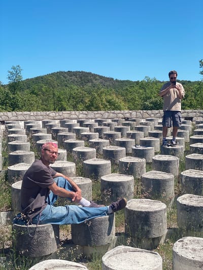 Two men are present in an outdoor setting filled with rows of cylindrical concrete structures, possibly pillars or posts. The backdrop includes a hillside covered with trees and a clear blue sky.

One man, wearing a light-colored shirt and dark shorts, stands further away on the concrete structures, seemingly taking a photograph. He is wearing sunglasses.

The other man, closer to the foreground, is sitting on one of the concrete structures with his legs crossed. He is wearing a dark t-shirt and blue jeans, and also has pink hair.

The location may be Barutana, Montenegro.