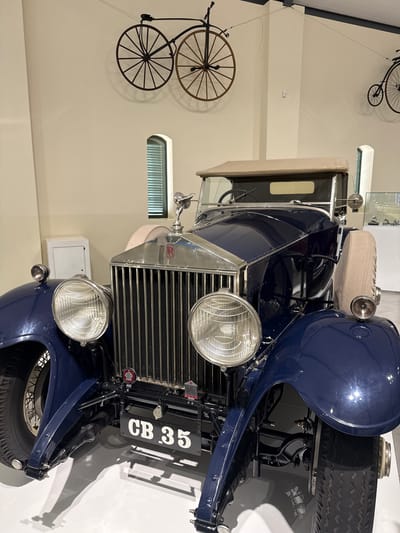 The image shows a vintage blue Rolls Royce automobile on display in a museum setting. The car's front grill, headlights, and license plate "CB 35" are prominent. Two penny-farthing bicycles are mounted on the wall behind the car. The image was taken indoors, likely within the Stellenbosch Local Municipality, South Africa. No people are visible.