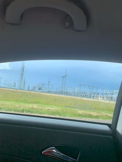 The image, taken from inside a vehicle, shows a view through the passenger window. The foreground is dominated by the dark interior of the car, including a door panel and a chrome handle. The window looks out onto a grassy field and a large electrical substation. The substation is comprised of numerous metal towers, insulators, and electrical wires, suggesting it is a high-voltage electricity transmission and distribution facility. The sky is cloudy and appears to be a light blue with some darker storm clouds in the distance. There are some faint reflections visible on the window, including what appears to be a Mercedes-Benz logo and some text which is difficult to decipher. The overall scene suggests a rural or semi-rural environment in the vicinity of the substation. The location of Курапольскі сельскі Савет, Belarus, is not explicitly visible in the image but this type of industrial infrastructure is common in such areas.