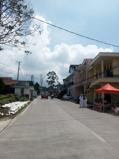 The image depicts a street scene in an urban or semi-urban area. The road is paved and stretches into the distance, flanked by buildings of various designs and colors. On the right side of the road, a red canopy is visible, possibly indicating a commercial establishment. Two individuals dressed in white garments are walking on the side of the road, away from the camera. A white car is parked nearby. Further down the road, a red vehicle is driving away from the viewer, followed by a motorcycle. Utility poles and wires run along the side of the road, and a few trees are scattered throughout the scene. On the left side of the road, there is a pile of white bags along the curb. The sky is partly cloudy.
The location cannot be confirmed to be in Zeyathiri District, Myanmar, based on visual elements alone.