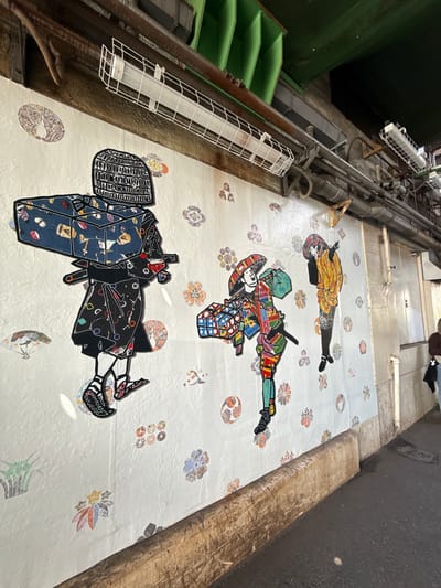 The image captures a floor in Shibuya, Japan, displaying three intricate figures or sculptures laid flat. The figures appear to be stylized human forms adorned with geometric patterns, cultural symbols, and vivid colors. They are arranged linearly on a light-colored surface patterned with smaller decorative motifs. The environment is industrial, possibly an interior space with metallic structures, pipes, and lighting fixtures visible on the left side of the image. The composition is shot from a skewed perspective, looking down at an angle towards the floor, adding a sense of depth and emphasizing the unusual art installation in this urban setting. A wooden wall is visible on the right.