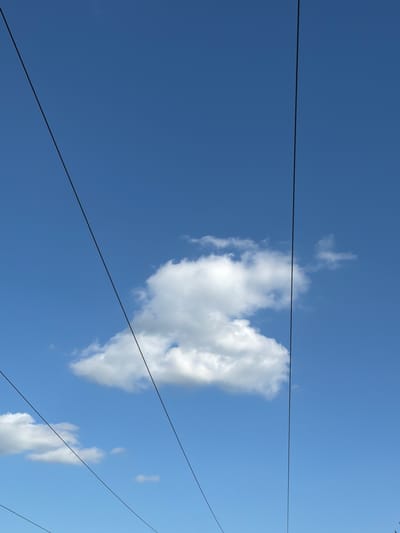The image captures a clear blue sky with several thin, dark wires stretching diagonally across the frame. One large, fluffy white cloud is centrally positioned in the upper half of the image. Two smaller clouds are visible towards the bottom left. The scene is likely an outdoor setting in Burnaby, Canada, possibly depicting power lines or a cable system against the sky. There are no people or discernible actions visible in the image.