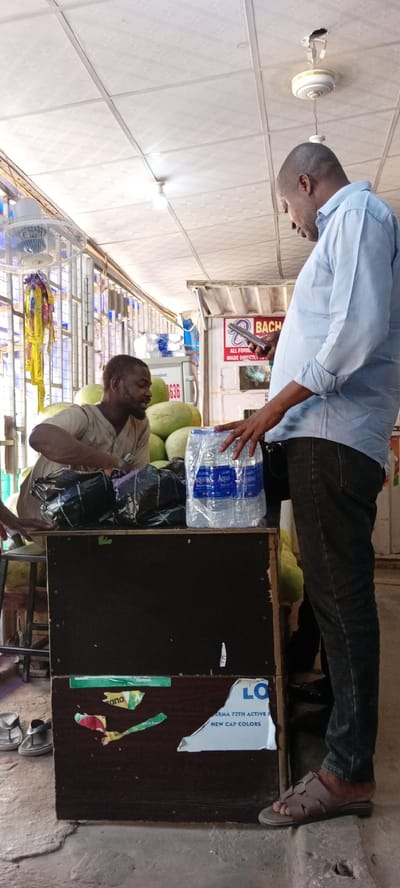 In Aviation Quarters, Nigeria, two men are interacting at a market stall. The man on the left, wearing a light brown tunic, is seated behind a dark wooden counter, handling a stack of black plastic-wrapped items. He appears to be looking towards the man on the right. The man on the right, wearing a light blue long-sleeved shirt and dark pants, is standing and looking down at the counter. He is holding a smartphone in his right hand and has his left hand resting on a plastic-wrapped package of Aquafina bottled water. The counter displays various stickers and labels, including one with "LO" and "NEW CAP COLORS". Behind the seated man, several large green fruits, possibly watermelons, are visible, along with stacks of bottled water. The environment appears to be an indoor market with a drop ceiling, fluorescent lighting, and a ceiling fan.