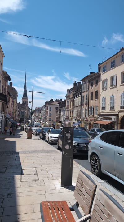 The image depicts a street scene in the city of Villefranche-sur-Saône, France, on a sunny day with a blue sky and scattered clouds. The street is lined with multi-story buildings featuring traditional architecture with shutters on the windows. Cars are parked along the right side of the street, and a few pedestrians are visible on the sidewalk on the left. In the distance, a tall church spire dominates the skyline. Two wooden benches are in the foreground on the right, facing the street. A lamppost and a trash bin are also present on the sidewalk. A sign with text "Circuit des Trésors cachés" is affixed to a metal pole in the middle ground. The pavement is made of stone tiles.