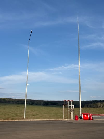 The image shows two vertical metal poles against a blue sky with wispy clouds. One pole is topped with what appears to be a small white rectangular object, while the other has a dark, rectangular object at its top. The bottom of the image includes a narrow strip of asphalt, a grassy area, and a line of trees on the horizon. Adjacent to the road, there are what look like metal pipes with red caps and a red equipment cart. There are no people visible. Based on the information, the location is Ermolaevo, Russia. The scene appears to be an industrial or infrastructural area.