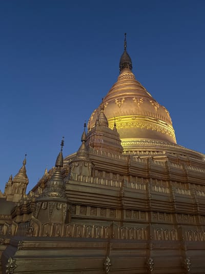A golden Burmese stupa rises against a deep blue sky. The bulbous main structure gleams with reflected light, intricately adorned with horizontal patterns. Smaller, tiered structures cascade down, featuring ornate detailing and spire-like tops. The perspective is looking upwards, highlighting the monumental scale and architectural complexity of the gilded temple.