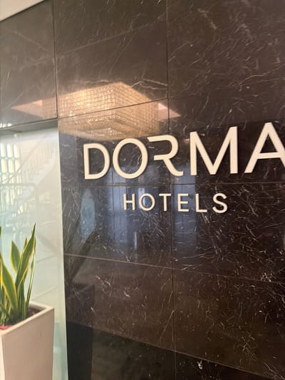 The image shows the exterior signage for Dorma Hotels, located in Espinho, Portugal. The sign, made of white letters against a dark marble-patterned wall, reads "DORMA HOTELS". To the left of the sign is a glass pane with a view of an indoor staircase and a potted snake plant. The overall scene suggests a modern hotel entrance.