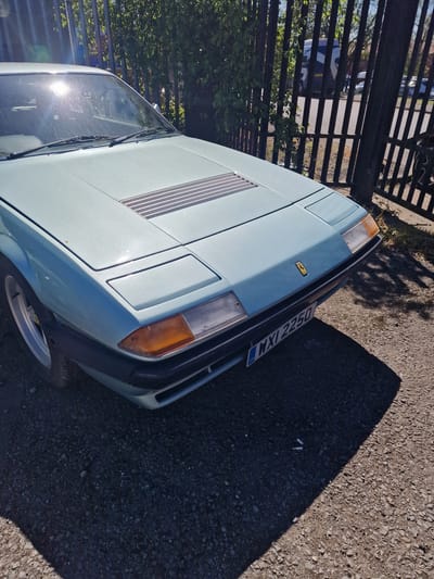 The media file displays a light blue classic Ferrari sports car, likely a 308 GT4, parked outdoors on a gravelly surface. The car is angled, showing its front-right side, including a pop-up headlight cover, an orange turn signal/side marker, and a visible portion of the front grille. A distinctive yellow rectangular Ferrari emblem is on the front hood, which also features narrow, horizontal vents or louvers. The car's windshield shows significant sun glare and reflections.
To the left of the car, a dark metal fence runs horizontally across the frame, with green foliage visible behind it. Strong, dark shadows are cast on the gravel in the foreground and to the right, indicating bright sunlight. The time of day appears to be mid-day or early afternoon due to the harsh lighting and distinct shadows. The weather is clear and sunny.
The car bears a UK license plate, clearly displaying "GB" on the left and the registration "WYL 2250." This, combined with the provided context, places the scene in South Tyneside, United Kingdom. There are no people visible, and the car is stationary, suggesting it is simply parked. Two small white cylindrical objects, possibly cigarette butts, are visible on the shadowed ground area in one of the crop images.