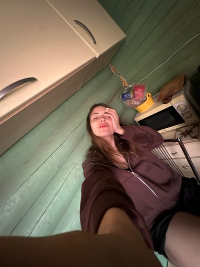 A young woman with long brown hair is shown from the chest up, taking a selfie in what appears to be a kitchen. She is wearing a dark maroon zip-up hoodie and black shorts. Her left hand is raised to her forehead, and her eyes are closed. The background shows wooden walls painted a light green, a beige cabinet with silver handles, and a microwave on a shelf. A yellow basket with colorful plastic cups is also visible. The image was taken in Zhebrei, Russia.