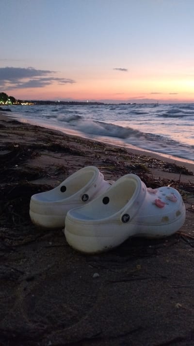 Two white Crocs are placed side-by-side on a beach in Neoi Epivates, Greece, as the sun sets over the Aegean Sea. The sky displays a gradient of blues, pinks, and oranges, with scattered clouds. The sea is calm with gentle waves lapping the shore, which is covered in sand and seaweed. In the distance, the lights of the town are visible, hinting at the presence of a settlement along the coastline. The image captures a serene moment of relaxation and the beauty of a Mediterranean sunset.