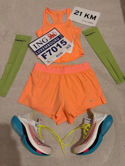 The image shows running apparel and accessories laid out on a textured surface. There are two pairs of light green arm sleeves with "PENALTY." printed on them. Next to them is a white sign with "21 KM" printed in black, along with the words "ING Luxembourg". There is an orange Nike running top and a pair of orange running shorts also from Nike. A bib number is placed on top of the top, marked "ING Luxembourg 20" with the number "F7015" and the name "ROSI". Lastly, there are two Saucony brand running shoes with light blue soles and yellow laces.