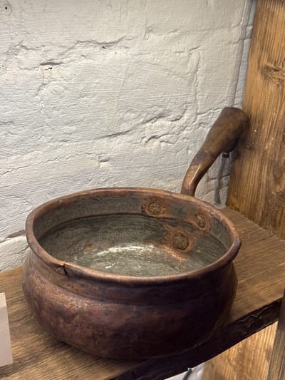 An antique copper cooking pot with a curved handle hangs on a wall. The pot is tarnished, with visible corrosion and dark spots. Three circular indentations with material inside are visible on its side. The pot is positioned against a textured white wall and a weathered wooden beam. Another wooden beam is visible on the right, with a dark metal object leaning against it. The scene is indoors, with a rustic aesthetic. There is no visible evidence indicating the city of Novo-Nikol'skoe, Russia, in the image.