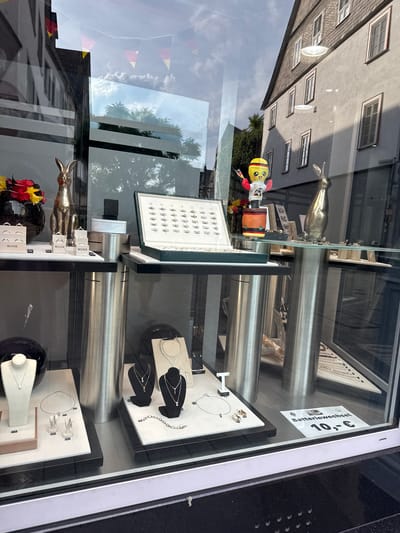 The image shows a street-level view of a jewelry store's window display in Wetzlar, Germany. The display features a variety of jewelry items, including necklaces, earrings, and rings, arranged on white and black pedestals and in display boxes. Notably, there are two metallic rabbit figurines, one gold-colored and the other a lighter silver, positioned prominently. A small, animated doll wearing a yellow shirt with a soccer ball design and holding German flags stands on a cylindrical object. German flag bunting is visible reflected in the window from above. In the background, the facade of a building with multiple windows and a tiled roof is visible, along with trees and the sky. A sign in the lower right corner of the window reads "Batteriewechsel 10,- €", indicating a battery replacement service. The overall scene suggests a commercial establishment presenting its merchandise to the public.