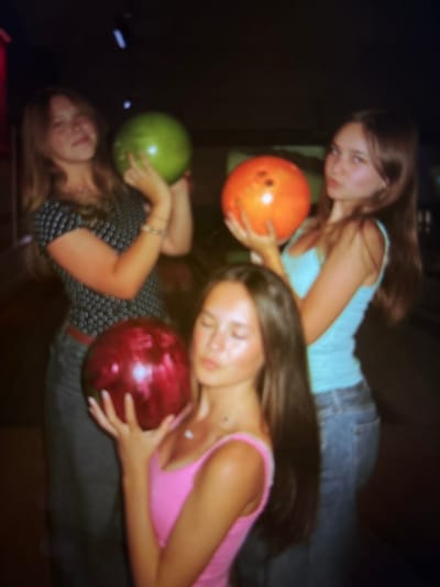 The image depicts three young women in a bowling alley. The woman on the left, with long brown hair, is holding a green bowling ball. She is wearing a dark patterned shirt. In the center, another woman with long brown hair is holding a red bowling ball and wearing a pink tank top. The woman on the right, also with long brown hair, is holding an orange bowling ball and wearing a light blue tank top and jeans. The background is dark and out of focus, with some blurry lights suggesting the environment of a bowling alley. The lighting is dim and appears to be from artificial sources within the bowling alley. There are no visible landmarks or indicators that would suggest the specific location within the city of Pori, Finland.