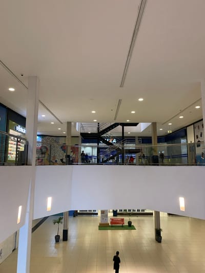 The image displays the interior common area of Shopping Camará, a multi-level commercial mall located in Camaragibe, Brazil. The perspective is from an upper floor, providing an expansive view of the ground level atrium and an adjacent mid-level.
The environment is characterized by modern architecture, featuring a high, white ceiling with integrated circular recessed lighting fixtures and linear air conditioning vents. Large white, rectangular support columns and broad white wall structures dominate the space. The ground floor is paved with light-colored square tiles, arranged in a grid pattern. Upper levels are delineated by transparent barriers topped with stainless steel handrails.
On the ground floor, a single individual, dressed in dark clothing, is observed walking away from the camera towards the rear of the atrium. Centrally located on this floor is a red slatted bench positioned on a square green mat, flanked by two potted plants in dark square planters. A vertical sign structure is visible behind this seating arrangement.
The mid-level, located directly across from the viewer, features several commercial establishments. To the left, a storefront clearly displays the "o Boticário" logo, showcasing products behind a glass facade. To the right, another commercial space with blue wall accents is partially visible. In the background of this level, a prominent black metal staircase with multiple sections and metal handrails connects to higher floors. Approximately 10-15 individuals are dispersed across this mid-level and the floor above it, engaged in walking or standing near storefronts and railings, indicating general public circulation. One storefront on the mid-level displays a large, colorful abstract mural. A small dome-shaped security camera is affixed to the ceiling near the "o Boticário" store, and a green "EXIT" sign is visible above a distant doorway on an upper level. Vertical light apertures are integrated into the lower sections of the white wall structures on the left and right.