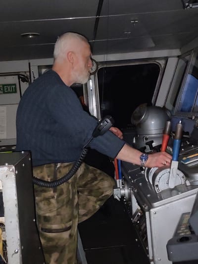 A man stands in what appears to be the control room of a ship or vessel, operating a control panel. He is Caucasian, with short white hair and a white beard. He wears a dark blue sweater and camouflage patterned pants. His left leg is partially visible, appearing to be elevated. He is holding a microphone in his left hand and manipulating a lever on the control panel with his right. He is wearing a watch on his left wrist. The control panel is metal and features multiple levers and controls. Behind the man is a window showing a dark view of the outside. An "EXIT" sign is visible on a wall to the left. The location cannot be determined as being in the city of Uzhla, Russia based on the provided visual information.