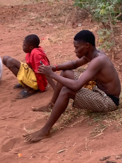 Two young, dark-skinned individuals are outdoors on a dirt ground. One person, shirtless and wearing patterned shorts, leans over another who is clad in a red shirt and yellow shorts. The shirtless person appears to be attending to the other, with their hands on the individual's back. The person in the red shirt is wearing one sandal. Grass and foliage are visible in the background.