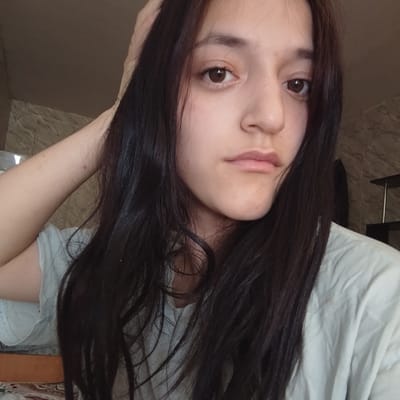 The image shows a close-up, waist-up shot of a young woman with long, dark hair. She is wearing a light-colored t-shirt. Her right arm is raised and her hand is resting on the back of her head. She has brown eyes and a neutral expression. The background is softly blurred, but appears to be an indoor setting with muted colors and textures on the wall. There is no visible indication of the city of Baymak, Russia.