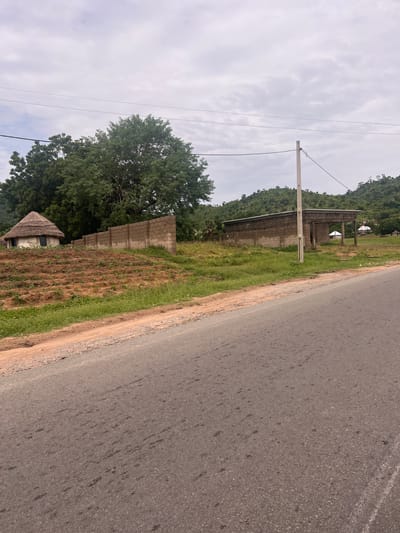 The image depicts a scene in Minda, Nigeria. The foreground features a paved road running diagonally across the frame, with a dirt shoulder and sparse vegetation. To the left of the road, a small, thatched-roof hut sits in a field of dry grass and young plants. Behind the hut, a wall constructed of concrete blocks separates the field from an area with lush green trees. Further in the background, there are more concrete block structures, possibly under construction or unfinished, and a densely forested hillside. Overhead, a cloudy sky dominates the upper portion of the image, with power lines stretching across it. The lighting suggests it is daytime, possibly overcast. The overall impression is of a rural or semi-rural environment with developing infrastructure. There are no visible people or text in the image.