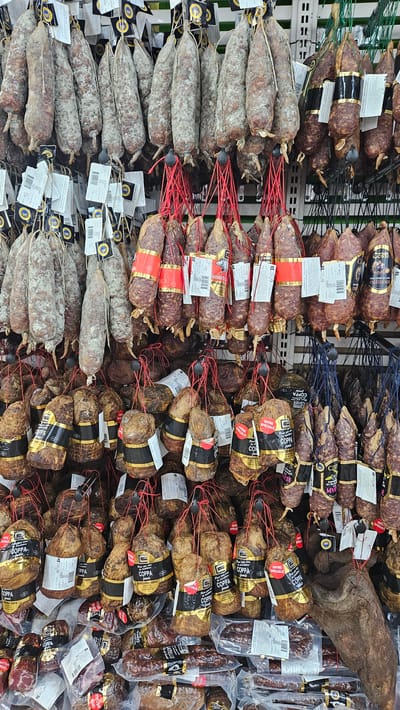The image displays a large assortment of cured meats, primarily sausages and hams, hanging from display racks in a market or shop. The meats vary in size, shape, and curing appearance, with some covered in white mold, others with dark, browned exteriors, and many wrapped in netting or butcher paper. Several items are tied with red twine. Labels are visible on many of the products, indicating various types of charcuterie and potential branding, some in French. The background consists of white metal shelving and what appears to be a green mesh or netting, suggesting a commercial display environment. The scene is well-lit, highlighting the textures and colors of the cured meats. The image is a still shot without any visible people. The location is identified as a shop in Corbara, France.