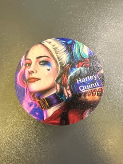 The image shows a round, circular object with a printed image of Harley Quinn, a fictional character. The image is depicted in a colorful, comic book style, with Harley Quinn's face prominently displayed. She has blonde hair with streaks of pink and blue, and her face is painted with blue and pink makeup, including a heart-shaped mark under her eye. The text "Harley Quinn" is printed in white letters on a blue background to the right of her face. The object appears to be made of wood, with visible wood grain. The object is placed on a textured, gray surface. There is no indication or context provided within the image that relates to Mikhailovka, Russia, or any specific city.