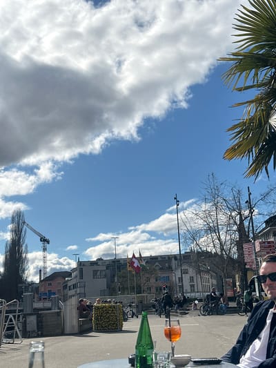 The image depicts a street scene in Schaffhausen, Switzerland, observed from an outdoor seating area, possibly a restaurant or café.

**Environment:** The sky is predominantly blue with scattered white clouds. The season appears to be spring, based on the presence of palm leaves in the upper left corner. The street is asphalt, flanked by buildings of varying heights and architectural styles. A construction crane is visible in the lower part of the scene.

**People and Actions:** A group of people ride bicycles in a peloton on the street. Patrons are sitting at the outdoor seating area, some obscured by the image's perspective. An individual wearing sunglasses and a jacket is partially visible on the right side of the frame.

**Objects:** A Swiss flag is displayed in front of a building. An aperol spritz, a bottle, and other glassware are present on the table in the foreground, suggesting a dining or social setting. Street signs are visible but unreadable. A yellow flower box sits along the edge of the sidewalk.