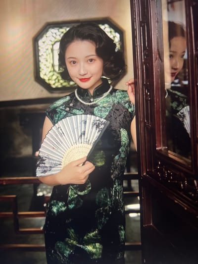 The media file features a young woman with dark hair styled in soft waves, her head tilted slightly to the side. She is adorned in a traditional Chinese qipao, a dark green dress with a floral pattern and a pearl necklace. In her hands, she holds a delicate fan, its surface decorated with a landscape painting and calligraphy. Her makeup is subtle, with rouged cheeks and a bright red lipstick. She appears to be posing for a photo, looking directly at the camera with a gentle smile.
The background suggests an indoor setting, possibly a traditional Chinese room or studio. A wooden frame, likely part of a piece of furniture or a mirror, is visible at the top of the frame. Through the frame, a reflection of the woman's face can be seen, adding depth to the image. The overall atmosphere is elegant and nostalgic, with hints of traditional Chinese culture. The lighting appears to be artificial, creating a warm and inviting ambiance.
The scene is set in Liandu District, China, though no specific weather or time of day cues are discernible from the image alone. The woman's expression is serene and poised. Visible text is present on the fan, written in Chinese characters, but it is not legible enough to be transcribed. The decorative elements of the qipao and the fan, along with the background, contribute to the overall traditional aesthetic.