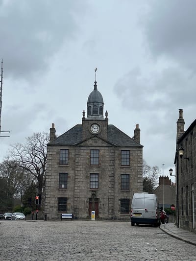 The image captures a view of the Tolbooth Museum in Aberdeen, Scotland. The prominent stone building features a clock tower topped with a dome and weather vane. The sky is overcast. A white van drives on the cobblestone road adjacent to the building, and a red traffic light is visible near bare tree branches. A street sign is attached to the building and parked cars line the street. The building's windows are dark. The surrounding buildings are also constructed of stone.