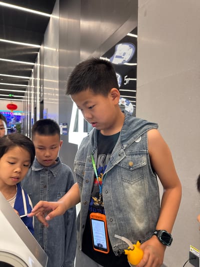 The scene is set indoors within a modern commercial or public building, specifically in Guangming District, China, indicated by the background elements. Three East Asian children are visible in the foreground, focused on a white, angled interactive display device. The central figure, an approximately 8-12 year old male, wears a faded denim sleeveless hooded vest over a dark t-shirt with a "KISS" band logo. He has a green lanyard around his neck, from which an orange-cased smartphone displaying text hangs. A black digital smartwatch is on his left wrist. He extends his right index finger to touch or point at the white display. In his right hand, he holds a yellow, possibly cartoon-themed, water bottle with a straw. To his immediate left, an approximately 6-10 year old male, dressed in a long-sleeved light blue denim shirt, intently observes the device. Further left, a roughly 5-9 year old female in a white garment and a blue and white striped top also gazes at the display. A fourth child's head is partially visible behind them on the far left. The background features a well-lit corridor with a dark ceiling containing linear lights, reflective metallic wall panels, and black display panels with white abstract graphics and a prominent "5F" indicating a floor level. A red decorative lantern hangs in the distant left background. The children are actively engaged in examining or operating the interactive device.