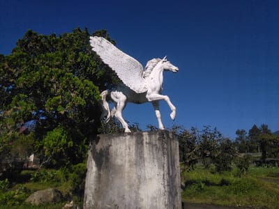 A white statue of a Pegasus is centrally positioned, rearing up with its front legs lifted and large, feathered wings spread upwards. The statue, appearing aged with some surface discoloration, rests on a grey, weathered concrete pedestal. The scene is set outdoors under a clear, bright blue sky in Batulawang, Indonesia. To the left and behind the statue, dense green foliage from large trees and bushes dominates, with hints of distant structures barely visible through the vegetation. To the right, an open grassy field with scattered smaller trees and shrubs extends into the background, leading to a distant, hazy tree line or hills. The lighting suggests direct sunlight. No individuals are present in the image.