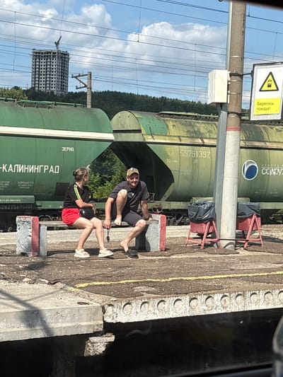 At a train station in Zelenodol'sk, Russia, two individuals are seated on a bench by a train track. On the left, a woman with blonde hair pulled back in a bun wears a black t-shirt with colored stripes and red shorts, with white sandals on her feet. She holds a dark purse in her lap. To her right, a man with a baseball cap on his head and a dark t-shirt and shorts is seated with his legs extended. Both appear to be waiting. Behind them, a long green train car, labeled "ЗЕРНО" (GRAIN) and "-КАЛИНИНГРАД", is parked on the tracks. Further in the background, a tall, unfinished concrete building is visible against a blue sky with scattered clouds, and a dense line of trees borders the horizon. A utility pole with a sign warning to "Beware of the train!" stands to the right of the individuals, with two red trash receptacles with black bags placed at its base. The ground is a weathered concrete platform.
