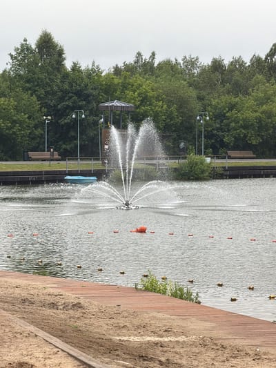 The image depicts a calm lake with a fountain in the center. Along the shore, there is a sandy beach area with a wooden walkway. Trees and a clear sky form the background. Several orange buoys are floating in the water, marking a boundary. In the distance, there are benches and lampposts, suggesting a park or recreational area. A small blue boat is visible near the shore. The overall impression is that of a peaceful, outdoor environment.