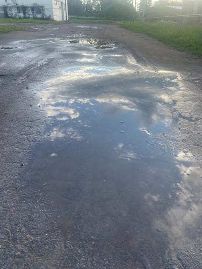 The image shows a cracked asphalt surface with multiple puddles of water. The largest puddle in the foreground reflects a blue sky with white, puffy clouds and sunlight. To the left, in the background, is a weathered white building with some visible piping. The environment appears to be an outdoor area, possibly a yard or industrial space, with vegetation and a dirt path or road visible further in the background. The overall impression is one of a somewhat neglected urban or industrial landscape after rain.