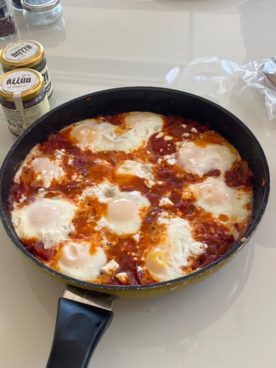 A black frying pan, with a yellow-gold underside and a black plastic handle, is centered on a reflective, light-colored surface. The pan contains a dish identified as shakshuka, featuring eight distinct, partially submerged fried eggs with firm white yolks and bright orange centers, nestled within a thick, chunky reddish-orange tomato sauce. The sauce shows visible pieces of tomato and scattered white crumbles, consistent with feta cheese. In the upper-left background, two small glass jars with gold lids are present, displaying Hebrew text on their labels, including the brand "עליא" (Alia) and the product "אגוזים" (walnuts). A taller clear jar with a silver lid and a smaller glass container with red flakes are also visible behind them. To the upper-right, a crumpled transparent plastic wrap lies on the surface. The scene is brightly and evenly lit. This event is located within the Merom HaGalil Regional Council, Israel.