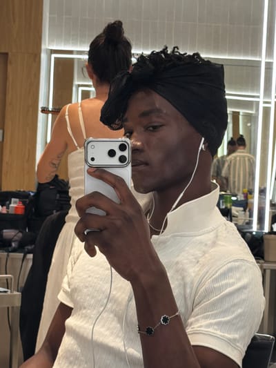 A young Black person with dark skin and curly hair is taking a selfie in a well-lit room, possibly a salon or dressing room, with wooden paneling and bright strip lighting visible in the background. They are wearing a black headscarf, a white collared shirt with a textured pattern, and white earbuds. A silver bracelet with four black clover-shaped charms adorns their left wrist. They are holding a silver smartphone in their right hand, which is pointed towards the mirror. In the reflection of the mirror, another person with dark hair in a bun is visible from behind, wearing a white top with spaghetti straps. Further in the background, other people are also reflected in the mirrors. Various items, possibly makeup or hair products, are arranged on shelves in the background. The overall impression is that of a behind-the-scenes or preparation area.