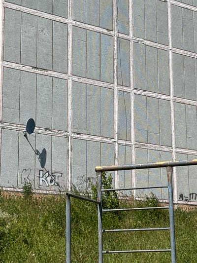 The image shows a section of a weathered building facade with a grid pattern of glass panes, possibly frosted or textured, set within light-colored frames. There is some graffiti on the lower portion of the building, including the letters "K KOT" and other illegible markings. In front of the building, there is a metal railing structure, suggesting a public area or park. Tall, green grass and some bushes are growing in front of the railing. A satellite dish is mounted on the building, casting a shadow. The scene appears to be outdoors, under daylight. The location is identified as Шолохово, Russia.